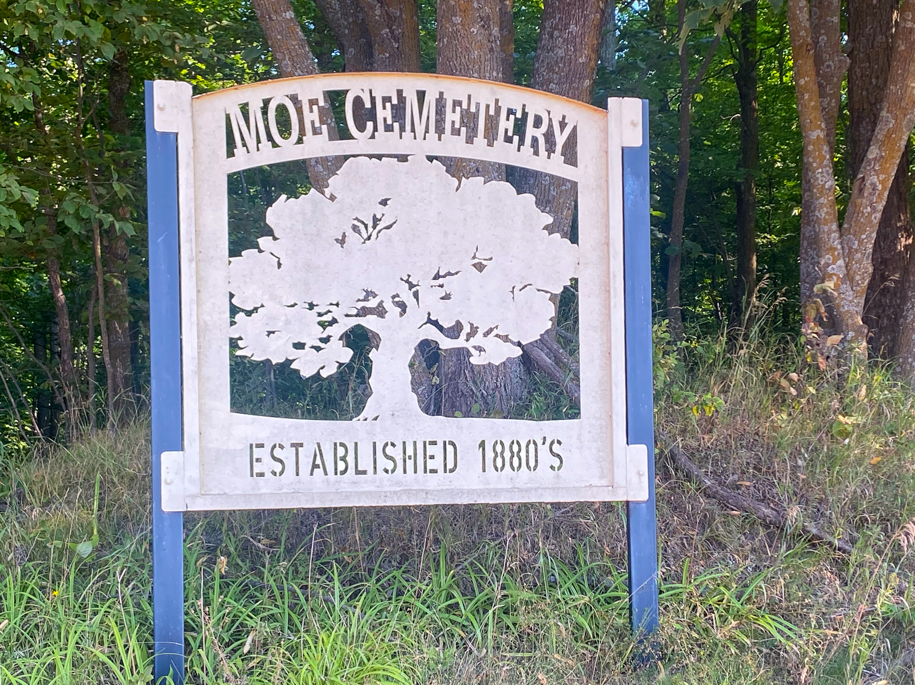 Outdoor sign reading “MOE CEMETERY” and “ESTABLISHED 1880’S” with a carved map/emblem and trees behind it.